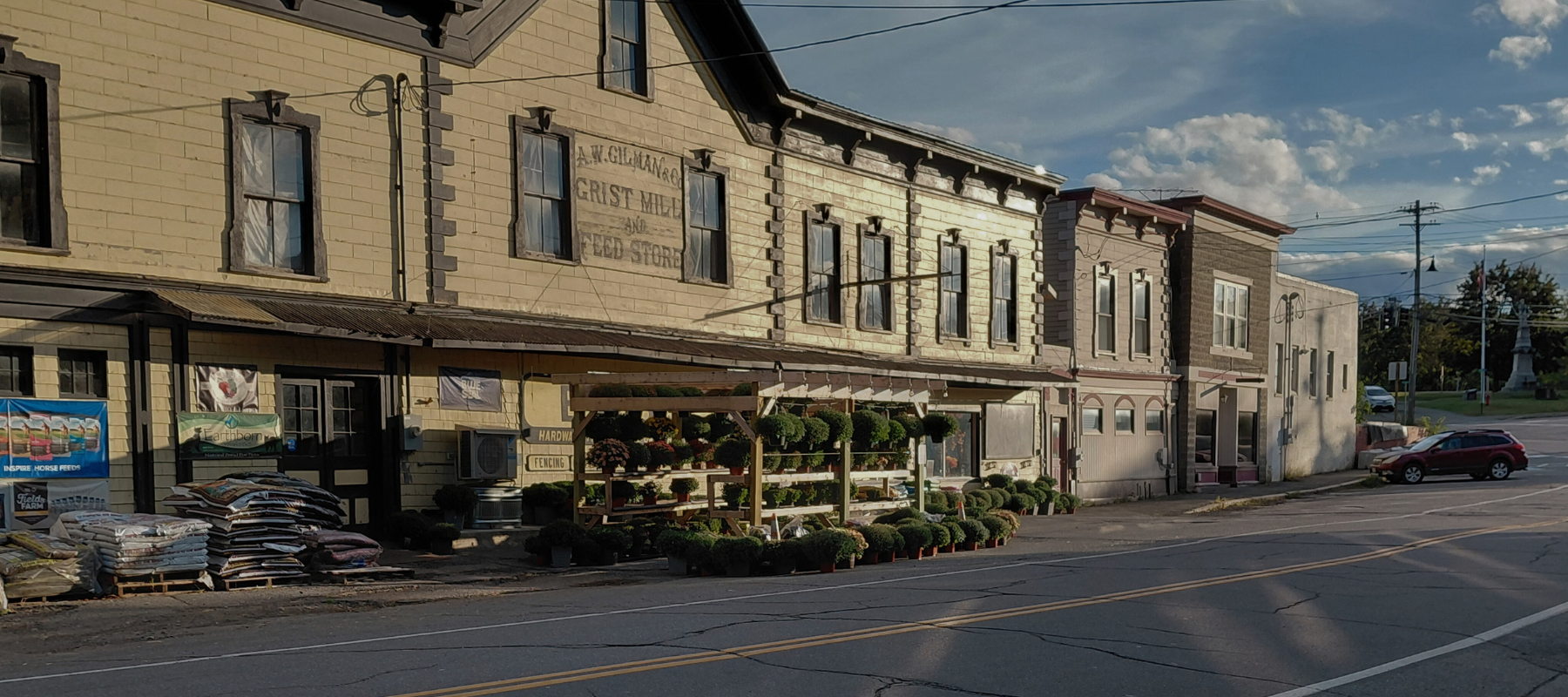 Historic wooden building on a sunny day with a rustic storefront displaying potted plants and soil bags. A red car is parked on the quiet street.